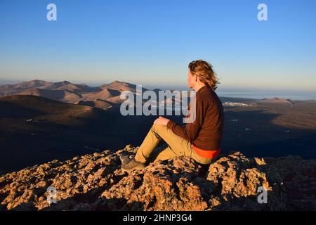 Sonnenaufgang auf Lanzarote. Eine Frau, die auf einem Berg sitzt und die vulkanische Landschaft mit den kleinen Städten Yaiza und Uga überblickt. Kanarische Inseln, Spanien. Stockfoto