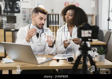 Multiethnische Ärzte mit männlichen und weiblichen Ärzten, die Videos aufzeichnen, während sie zusammen am Schreibtisch sitzen und über Pillen sprechen. Zwei medizinische Mitarbeiter teilen sich mit Rat und Erfahrung in sozialen Netzwerken. Stockfoto