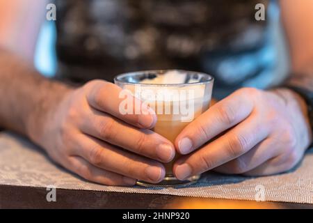 Konzentrieren Sie sich im Vordergrund auf die Hände eines Mannes, der ein Glas Cappuccino hält. Stockfoto