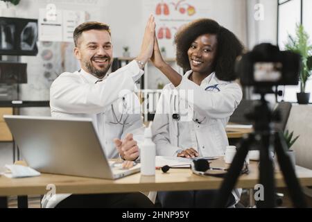 Positive, vielfältige medizinische Mitarbeiter geben sich fünf hoch, während sie am Schreibtisch sitzen und Videos auf einer professionellen Kamera aufzeichnen. Erstellung von Inhalten für medizinische Blogs. Stockfoto