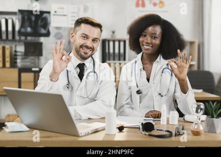 Zwei multiethnische medizinische Mitarbeiter, die mit einem modernen Laptop am Schreibtisch saßen, beschilderten das Schild und lächelten vor der Kamera. Konzept der Zusammenarbeit, des Erfolgs und der Wissenschaft. Stockfoto