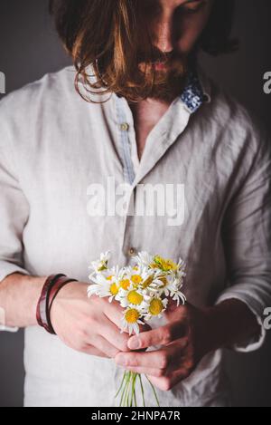 Bärtiger Mann in Leinenhemd mit langem Haar Holding Bouquet Von Gänseblümchen Blumen in den Händen Stockfoto