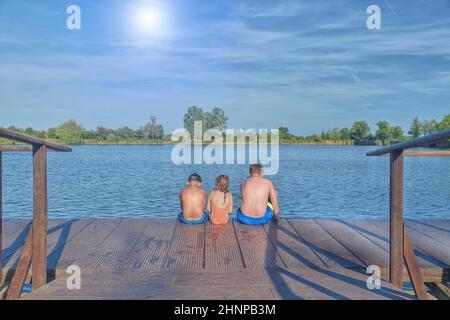 Kinder sitzen auf Pier. Drei Kinder verschiedenen Alters - jugendlicher Junge, elementare alter Junge und Mädchen sitzen auf einem hölzernen Pier. Sommer und Kindheit Konzept. Kinder auf der Bank am See. Geschwister sitzen auf hölzernen Pier Stockfoto