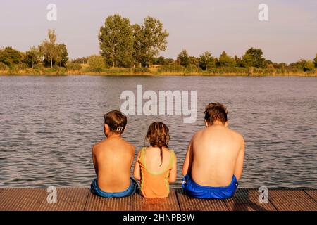 Kinder sitzen auf Pier. Drei Kinder verschiedenen Alters - jugendlicher Junge, elementare alter Junge und Mädchen sitzen auf einem hölzernen Pier. Sommer und Kindheit Konzept. Kinder auf der Bank am See Stockfoto