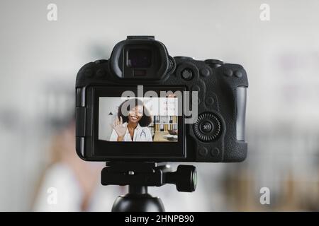 Videokamera-Bildschirm mit lächelnder afroamerikanischer Ärztin im weißen Laborkittel, die in einem Krankenhausbüro sitzt. Kompetente medizinische Mitarbeiterin, die Inhalte für ihren Kanal erstellt. Stockfoto