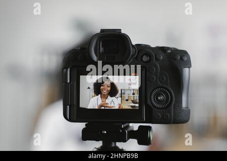 Betrachten Sie die positive weibliche Therapeutin, die während des Live-Streamings in der modernen Klinik über Pillen spricht, durch die Kamera. Afroamerikanische Frau im Laborkittel, die am Schreibtisch sitzt. Stockfoto