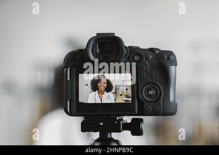 Videokamera-Bildschirm mit lächelnder afroamerikanischer Ärztin im weißen Laborkittel, die in einem Krankenhausbüro sitzt. Kompetente medizinische Mitarbeiterin, die Inhalte für ihren Kanal erstellt. Stockfoto