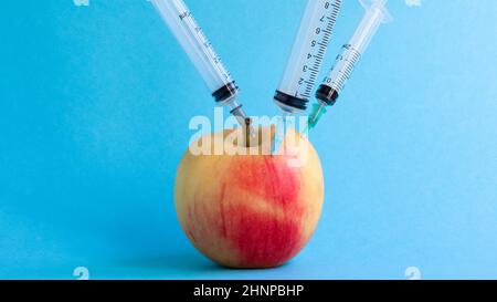 Drei verschiedene medizinische Spritzen, die auf blauem Grund tief in einen Apfel stecken. Die Frucht ist mit Spritzen bestreut. Ein reifer roter Apfel, von allen Seiten von Spritzen durchbohrt. Gesundheitswesen oder Kosmetologie. Stockfoto