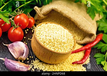 Roher Couscous in einer Schüssel und ein Sack mit Sackleinen, Knoblauch, Tomaten, Paprika und Kräutern auf dem Hintergrund eines dunklen Holzbretts Stockfoto