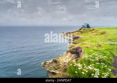 Eine Gruppe junger Menschen, die am Rand einer Klippe auf den berühmten Cliffs of Moher, Irland, sitzen Stockfoto