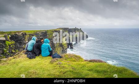 Eine Gruppe junger Menschen, die am Rand einer Klippe auf den berühmten Cliffs of Moher, Irland, sitzen Stockfoto