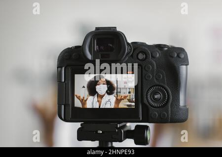 Videokamera-Bildschirm mit selbstbewusster afroamerikanischer Ärztin in weißem Laborkittel und Gesichtsmaske. Kompetenter Arzt spricht und gestikiert während der Videoaufzeichnung. Stockfoto