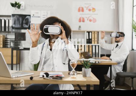 Die afroamerikanische Ärztin und ihre kaukasische Assistentin verwendeten VR-Brillen, während sie im Krankenhaus arbeiteten. Moderne innovative Technologie für medizinische Forscher. Stockfoto