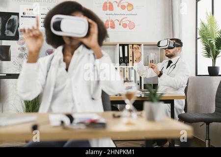 Zwei multikulturelle Ärzte mit VR-Brille, während sie in einem medizinischen Schrank sitzen. Kollegen, die beim Studieren des virtuellen Bildschirms in die Luft gehen. Stockfoto