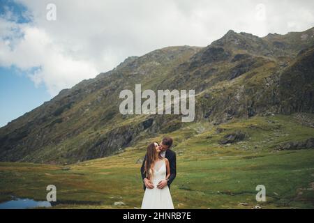 Portrait des Bräutigams umarmt und küsst die Braut von hinten durch die Schultern posiert auf Berge Hintergrund. Stilvolles Porträt. Glückliche Familie zusammen. Stockfoto