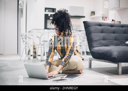 Junge Afro-Frau, die von zu Hause aus mit ihrem Laptop auf dem Boden sitzt Stockfoto