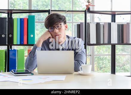 Ein junger, müde Geschäftsmann, der eine Brille trägt und den Kopf an der Hand vor dem Laptop am Schreibtisch hält, mit langweiligen und müden Gesichtschwehern Stockfoto