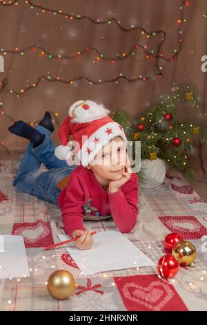 Weihnachtsjunge schreibt Brief an den Weihnachtsmann. Weihnachtshelfer Kinderbrief in rotem Hut Stockfoto