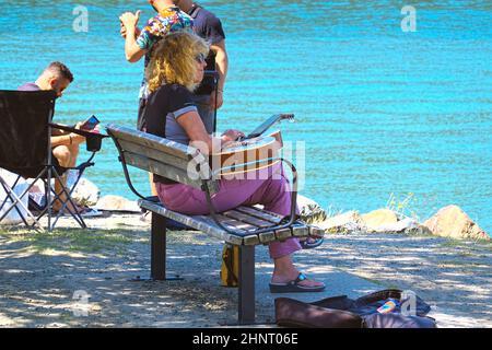 Blonde Frau in Sonnenbrille sitzt mit einer Gitarre auf einer Parkbank neben einem See. Stockfoto