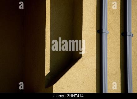 Formen und Muster aus Schatten, die durch das Sonnenlicht am Nachmittag auf die Außenfassade von zwei silbernen Metallausbrüchen auf gelbe Zement- oder Betonwände geworfen wurden Stockfoto