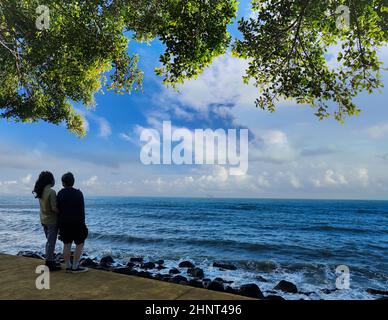 Ein paar junge Menschen, ein Mann in einer grauen Jacke und eine Frau in einer Jeansweste stehen an einem windigen Tag vor einem Hintergrund mit Blick auf das Meer Stockfoto