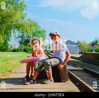 Entzückende kleine Mädchen und Junge auf einem Bahnhof, warten auf den Zug mit Vintage-Koffer. Reise-, Urlaub- und Kinderkonzept. Reiseversicherungskonzept Stockfoto