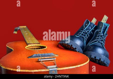 Rock und Roll Konzept. Schwarze Stiefel und akustischer Gitarre. Rock und Roll Stiefel auf Claret Hintergrund Stockfoto