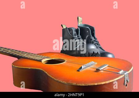 Rock und Roll Konzept. Schwarze Stiefel und akustischer Gitarre. Rock und Roll Stiefel auf rosa Hintergrund Stockfoto