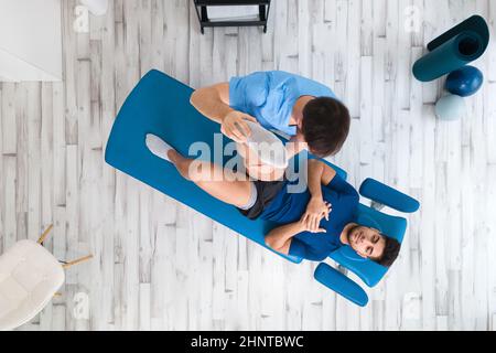 Ansicht von oben eines Therapeuten, der einem männlichen Patienten hilft, während er sein Bein im Krankenhaus streckt Stockfoto
