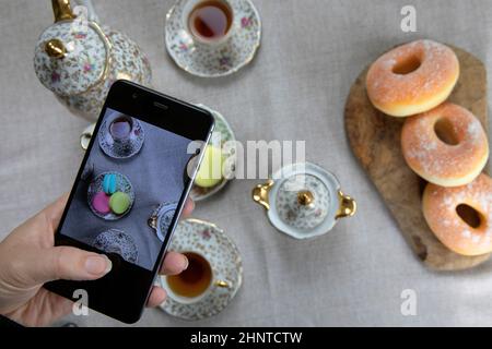 Draufsicht Bild der Dame, Blogger sitzen im Café und machen Foto mit Mobile von Lebensmitteln, Nachmittagstee mit Donuts und Makronen auf dem Tisch High ang Stockfoto