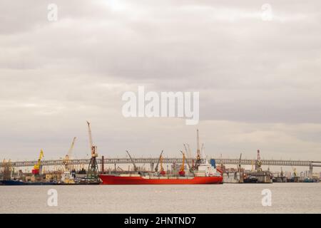 Rotes Frachtschiff im Hafen, Brücke über den Fluss mit Industriebetrieben, viele Industriekräne im Hafen Stockfoto