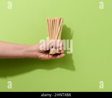 Weibliche Hand hält Einweg-Holzstäbchen zum Rühren von heißen Getränken auf grünem Hintergrund. Kaffee- und Teelöffel, kein Abfall Stockfoto
