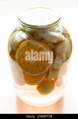 3-Liter-Glas mit hausgemachtem Kompot (geschmortes Obst) aus frisch geschnittenen Äpfeln und Orangen auf hellbraunem Tisch Stockfoto