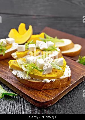 Bruschetta mit gebackenem Kürbis, gesalzenem Feta-Käse, Ricotta, Rucola und Gewürzen, Serviette und Gemüsescheiben auf dunklem Holzbrett-Hintergrund Stockfoto