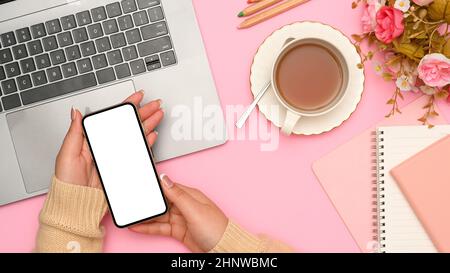 Oben Aufnahme einer weiblichen Hände, die ein Smartphone-Modell mit weißem Bildschirm über ihrem stilvollen rosafarbenen Schreibtisch hält. Laptop, Teetasse, Blumen und Schreibwaren auf Stockfoto