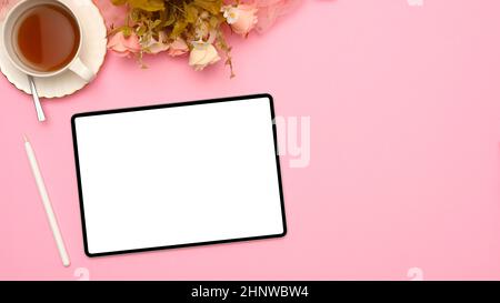Ein moderner rosafarbener Büroarbeitsplatz mit einem Mockup-Bildschirm aus digitalem Tablet-Touchpad, einer Tasse Tee und Blumen auf rosa Hintergrund. Stockfoto