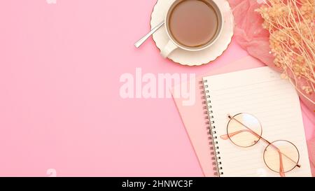 Ein minimalistischer femininer Arbeitsplatz im rosa Tischhintergrund mit Notizbuch, Brillen, Teetasse, getrockneten Blumen und Kopierraum. Draufsicht, flach liegend Stockfoto