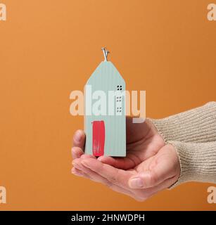 Zwei weibliche Hände halten ein Miniatur-Holzhaus auf braunem Hintergrund. Immobilienversicherungskonzept, Umweltschutz, Familienglück Stockfoto