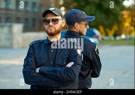 Zwei männliche Polizisten in Uniform und Sonnenbrille stehen Rücken an Rücken. Polizisten schützen das Gesetz. Bullen arbeiten auf Stadtstraßen, Ordnung und Gerechtigkeit Stockfoto