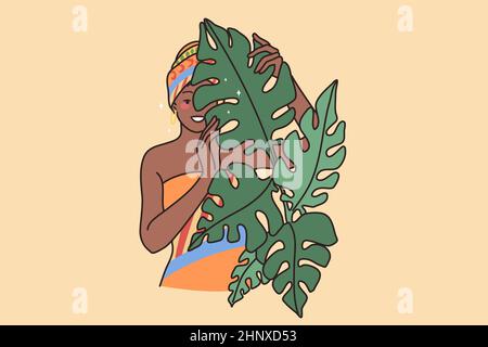 Junge lächelnde afroamerikanische Frau in traditioneller Kleidung und Turban. Glückliches schwarzes Mädchen mit Schal auf dem Kopf Blick hinter Blätter Bäume. Ethnische Zugehörigkeit und di Stockfoto
