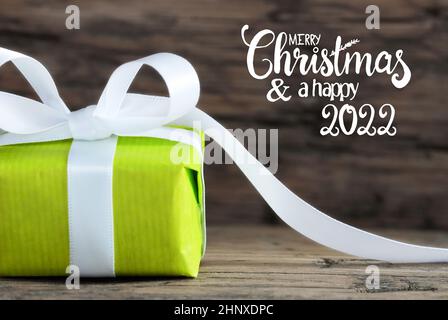 Label mit englischer Kalligraphie Frohe Weihnachten und Ein Happy 2022. Ein Grünes Geschenk Mit Weißer Schleife. Brauner Holzhintergrund Stockfoto