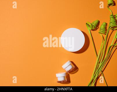 Leere weiße Glasgefäße mit Kunststoffdeckel auf orangefarbenem Hintergrund mit grünem Dekor. Verpackung für Creme, Gel, Serum, Werbung und Produktwerbung. Moc Stockfoto