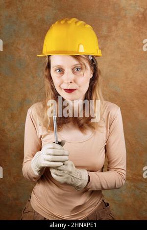 Junge Industriearbeiterin, die Schraubendreher hält und Schutzhelm und Handschuhe trägt Stockfoto