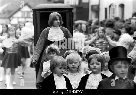 Die jährliche Knutsford Royal May Day Prozession im Jahr 1976 in Knutsford, Cheshire, England, Großbritannien. Traditionell umfasst es einen Kostümzug von Kindern in historischen oder legendären Kostümen mit Pferdekutschen. Hier gehen kleine Kinder in historischen Kostümen in der Prozession vor einem Teenager-Jungen, der die vorderen Griffe eines Wurf- oder Sedan-Stuhls trägt. Stockfoto