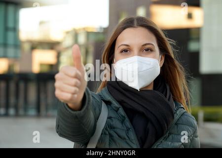 Junge optimistische Studentin gibt Daumen nach oben Geste. Sie trägt eine Schutzmaske (PSA), um Luftverschmutzung oder eine Coronavirus-Pandemie für saf zu vermeiden Stockfoto