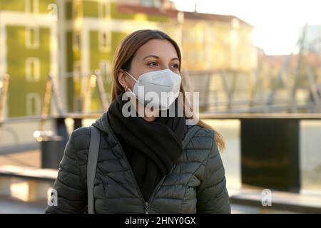 Schöne, lässige junge Frau mit einer schützenden KN95 FFP2-Maske, die am Sonnenuntergang in der Stadtstraße spazieren geht Stockfoto