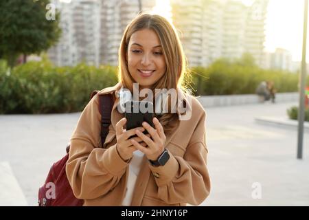 Sunshine Girl mit Mantel und Rucksack chattet mit Smartphone-App auf der modernen Stadtstraße bei Sonnenuntergang. Nachhaltiges Leben. Generation der Millennials. Headphon Stockfoto