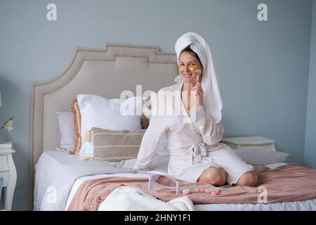 Glückliche Frau im Bett sitzend, Gesichtsmassage mit Jade-Gesichtswalze im Bademantel und Handtuch, natürliche Quarzsteine, entspannend, Zeit im Schlafzimmer zu Hause verbringen, in Nachdenklichkeit versinken, guten Tag haben Stockfoto