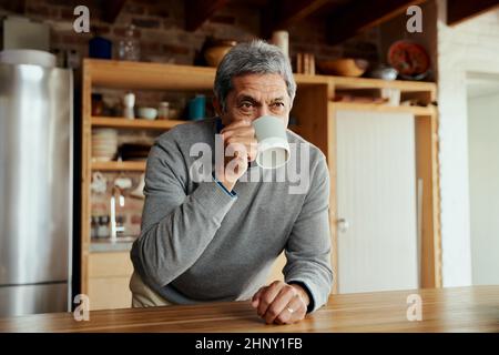 Porträt eines älteren Mannes im Ruhestand, der sich auf der modernen Küchentischtheke anlehnt und seinen morgendlichen Kaffee nippt. Stockfoto