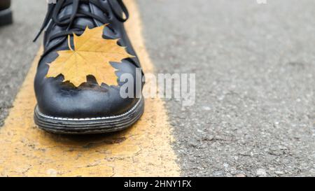 Beine in schwarzen Lederstiefeln auf dem Asphalt mit einem gelb gefallenen Ahornblatt. Herbstjahreskonzept, Herbstmode, trendy Lifestyle. Leerraum für Pos. Stockfoto